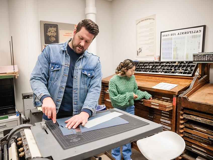 Students work in the Letterpress Studio in Communication Design. You can see the many slender drawers and cubbies which hold the individual letters to use in the Press.