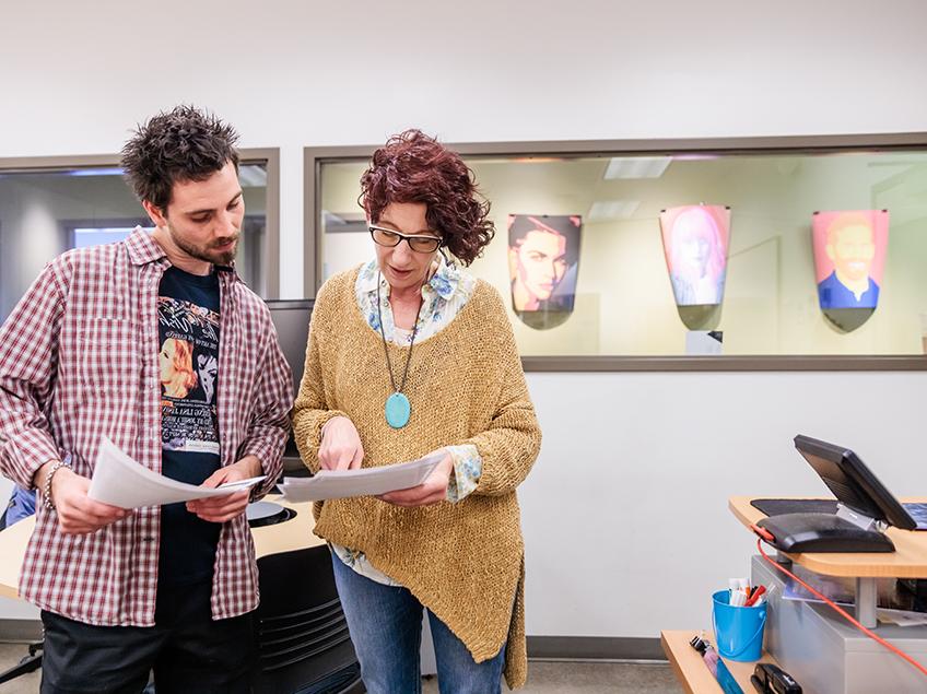 Professor Heidi Cies consults with a student about their final project. Posters on display in the background show graphics of larger-than-life portraits the students created.